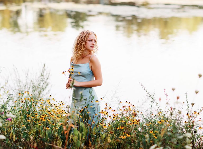 girl in blue dress with curly blonde hair standing near lake for senior pics